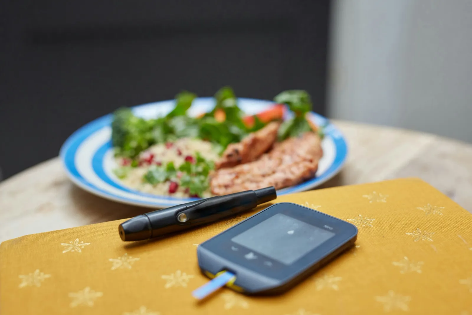 Glicosímetro e lancetador sobre a mesa, com um prato de salada, arroz e carne desfocado ao fundo