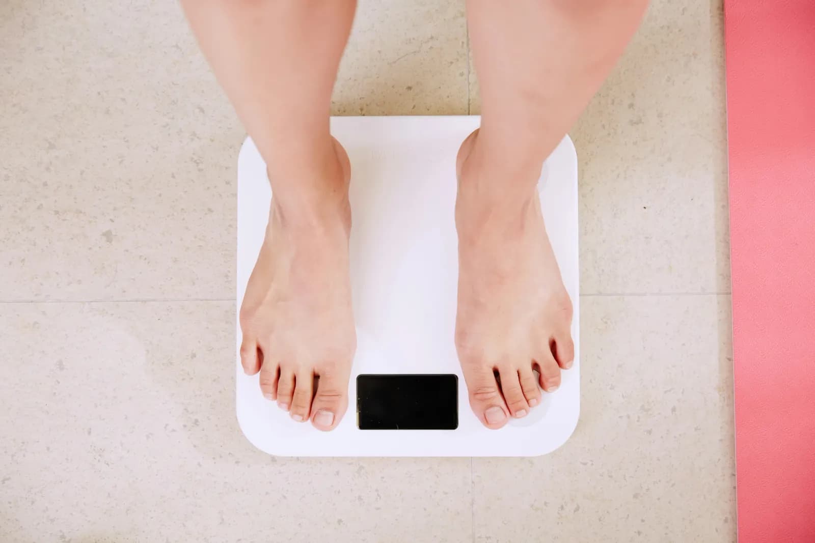 Pés descalços sobre uma balança digital branca, vistos de cima, ao lado de um tapete de exercício